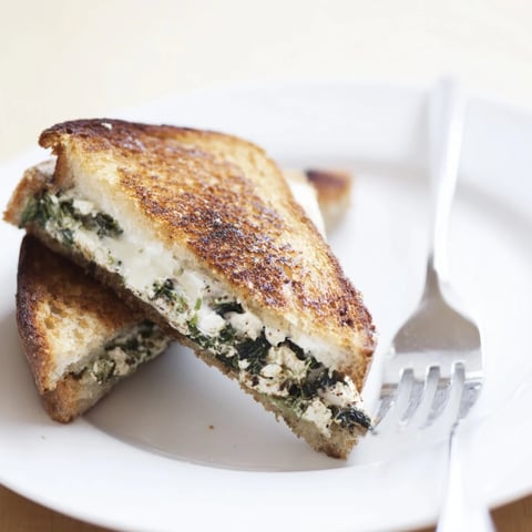 A close-up of Feta & Spinach Stuffed Grilled Cheese, golden-brown and melty, served on a rustic plate with fresh spinach leaves.