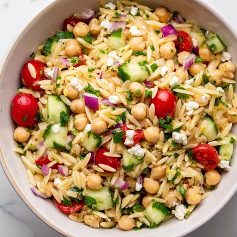 A vibrant bowl of chilled Lemony Chickpea Orzo Salad studded with herbs and chickpeas, ideal for a sunny picnic spread.  